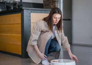 A woman kneeling while managing a robotic vacuum cleaner in a modern kitchen space.