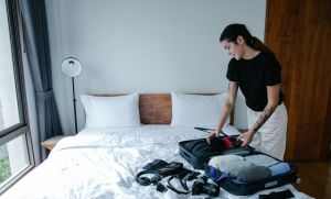 Woman packing suitcase with clothes and camera in a bright bedroom.