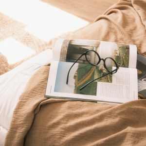A pair of eyeglasses resting on an open book on a cozy bed, basked in gentle sunlight.