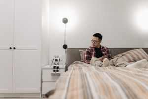A man in a modern bedroom sitting on the bed with a robot toy beside him, creating a tech-savvy atmosphere.